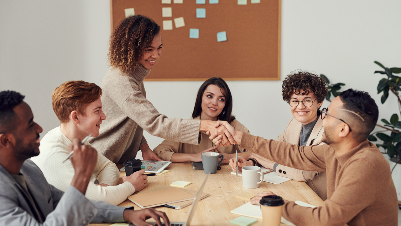 A smiling marketing team in a meeting to brainstorm for a campaign with two members shaking hands.